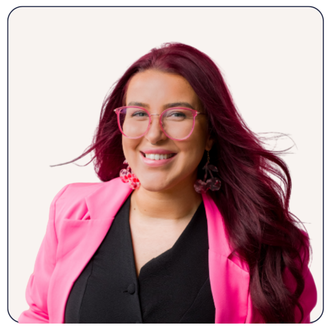 Head shot of woman with bright red hair smiling wearing pink glasses and a pink blazer.