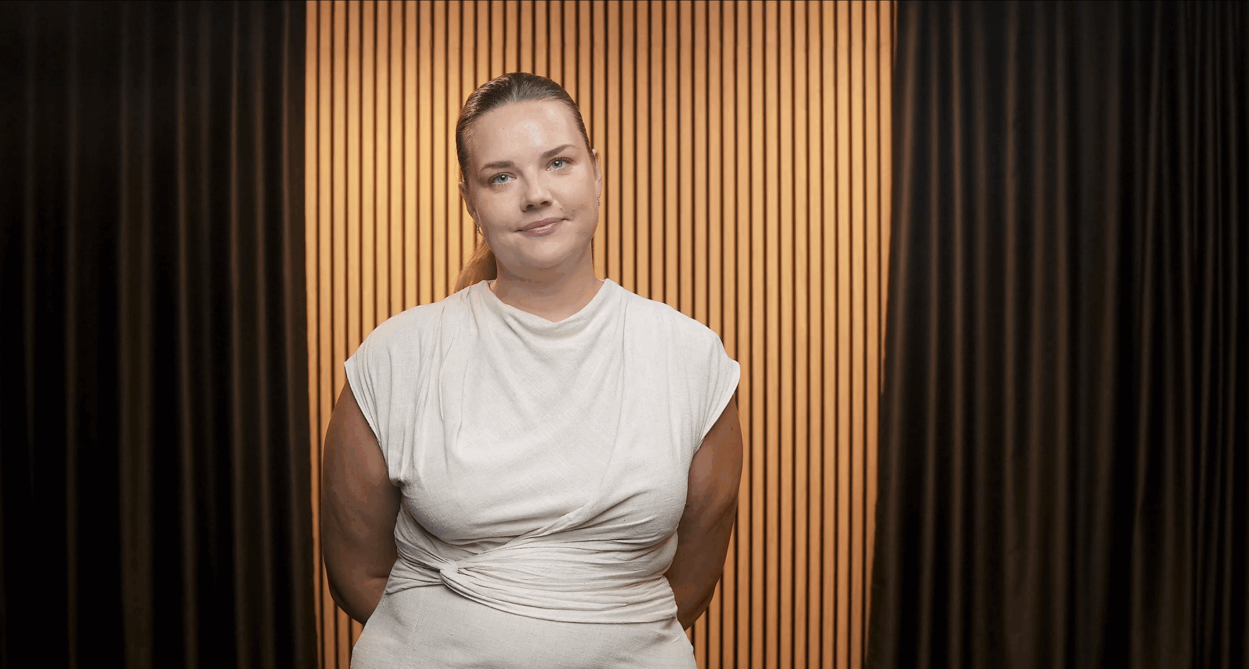 Headshot of a white woman with brown hair in a ponytail smiling at the camera stood in front of a background of slatted wood panelling