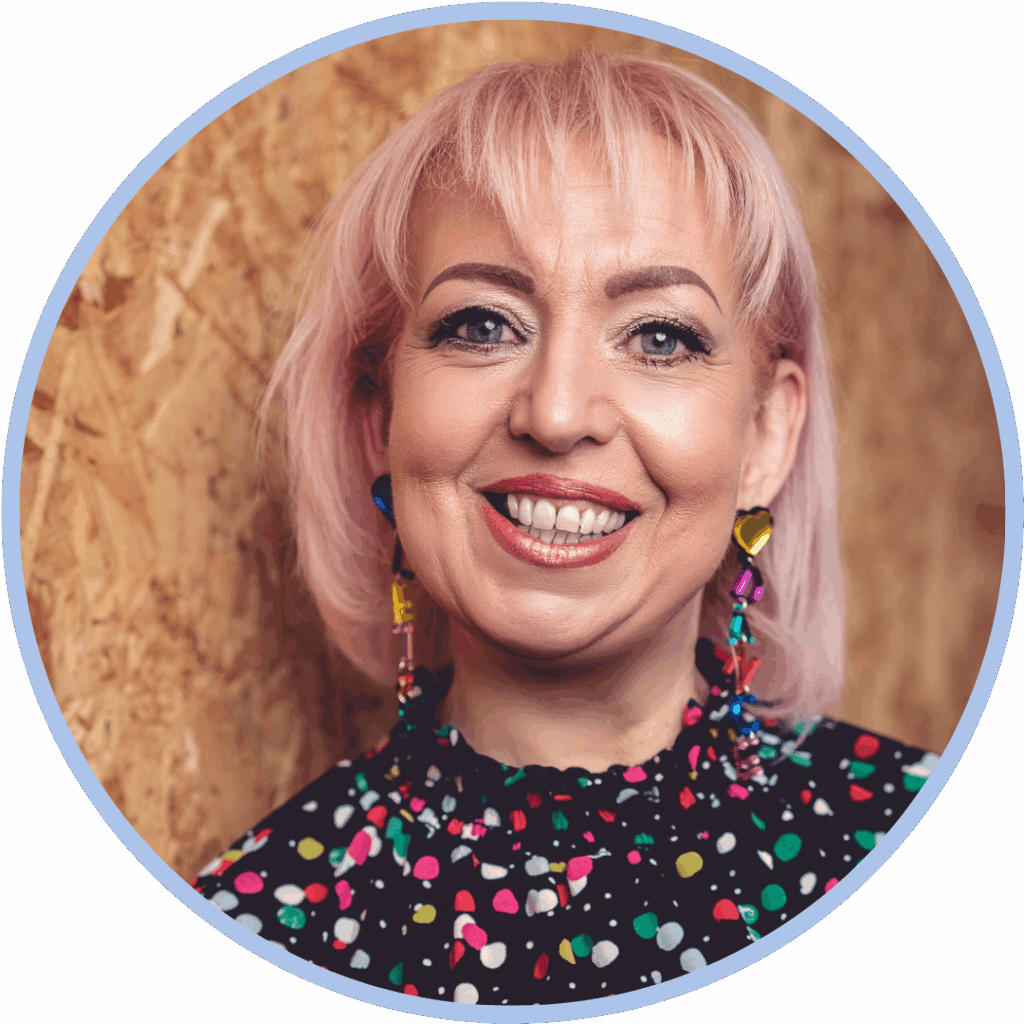 Headshot of a white woman with light pink hair wearing colourful earrings and smiling to camera