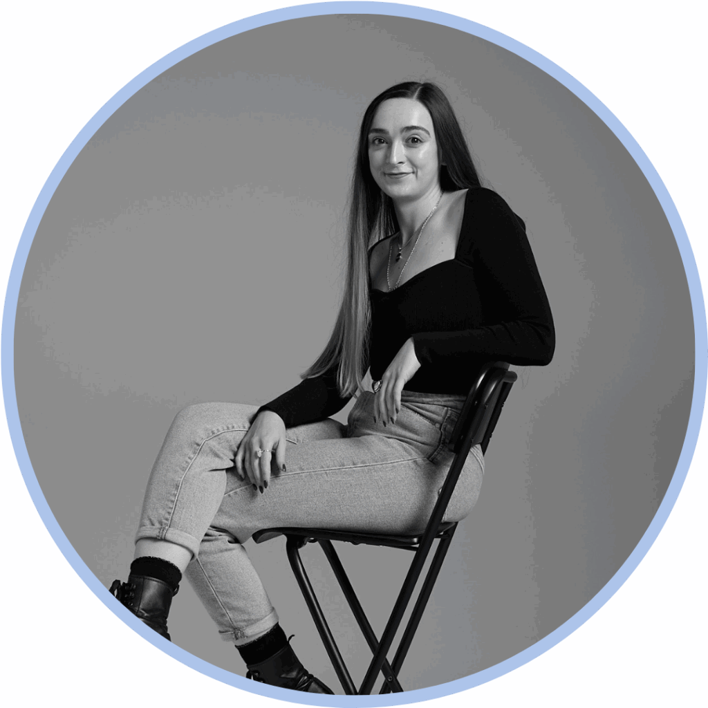 Black and white photo of a a white woman with long brown hair sat sideways on a directors chair leaning her arm on the back of the chair and smiling to camera