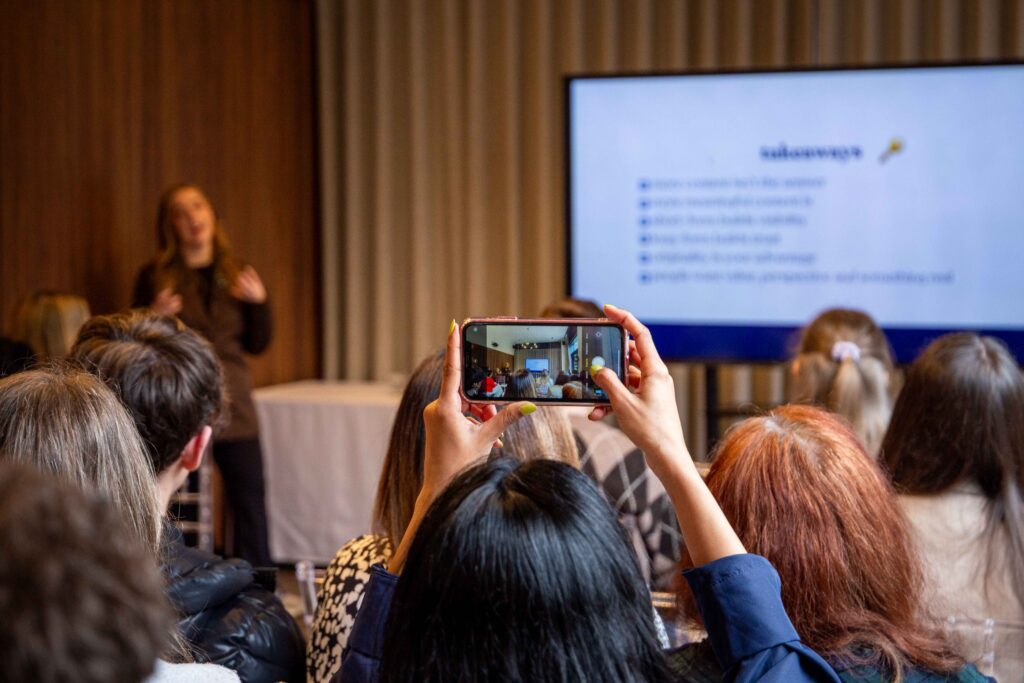Group of women sitting in a room taking photos on their phones and listening to a woman presenting